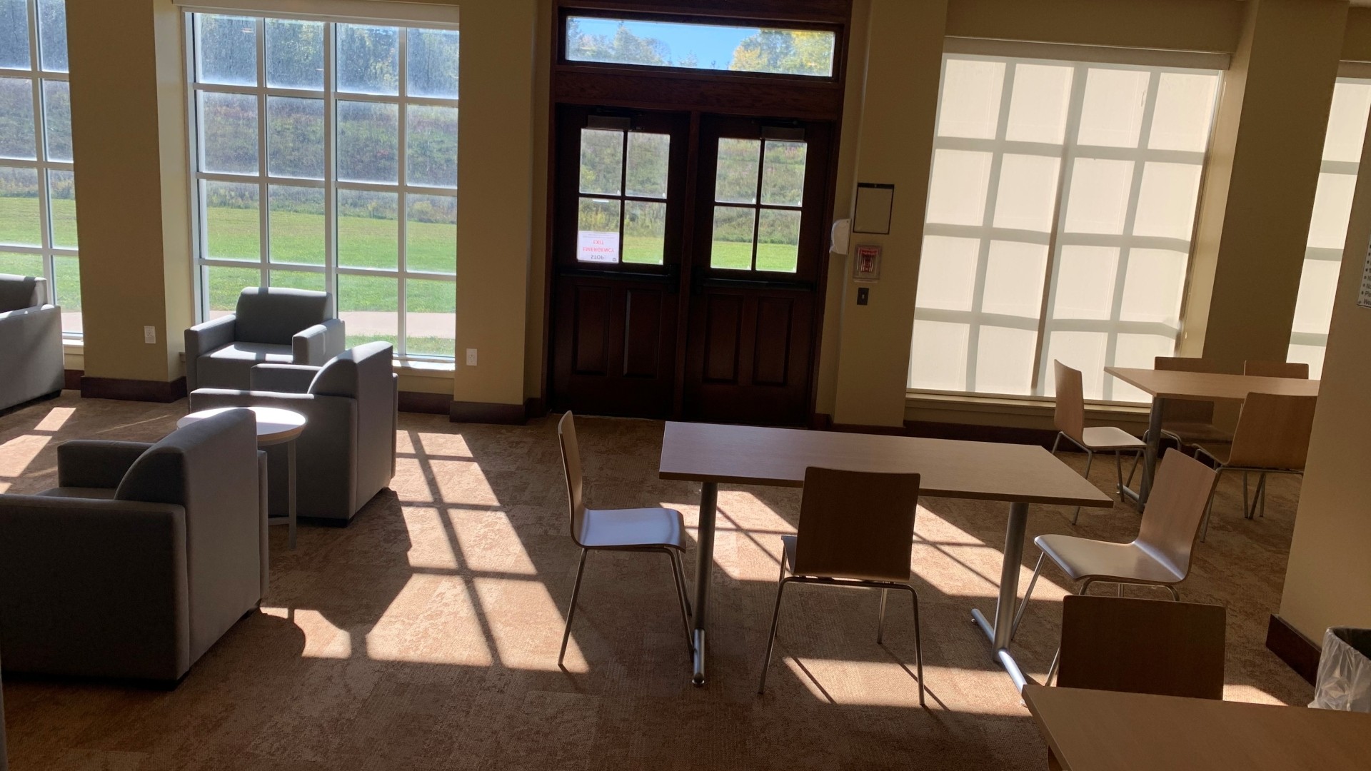 Lounge area with tables, chairs and large windows with the sunlight shinning through.