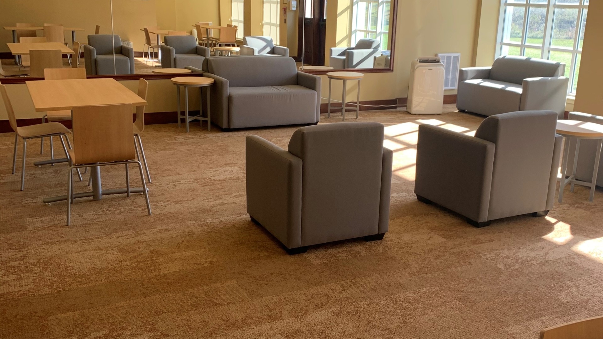 Lounge area with tables, chairs and large windows with the sunlight shinning through.