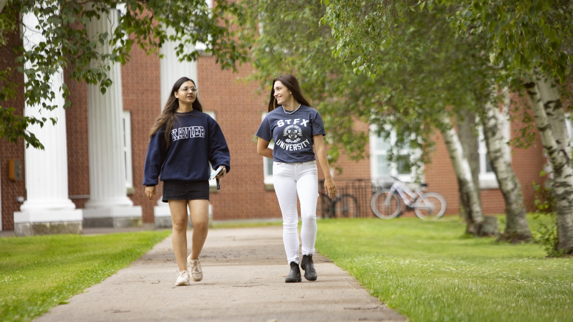 Students walking outside on a sunny day. There are trees around them and a bike rack in the background.
