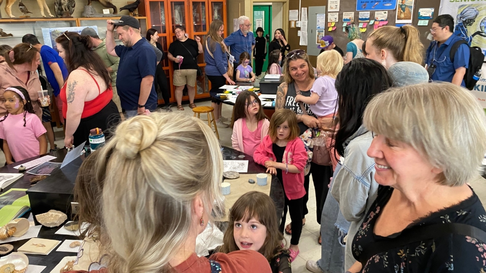 A crowded room with children and adults exploring tables with fossils and educational materials, surrounded by shelves with animal taxidermy.