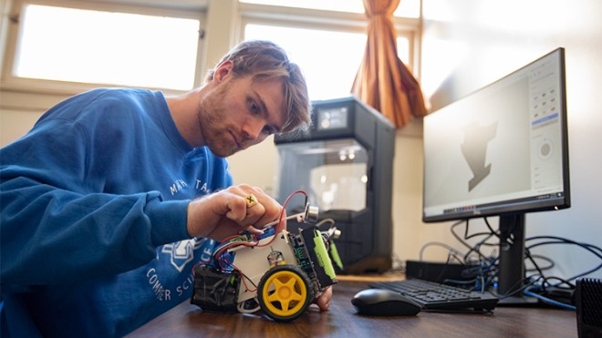 Person working on a small machine at a desk with a computer screen