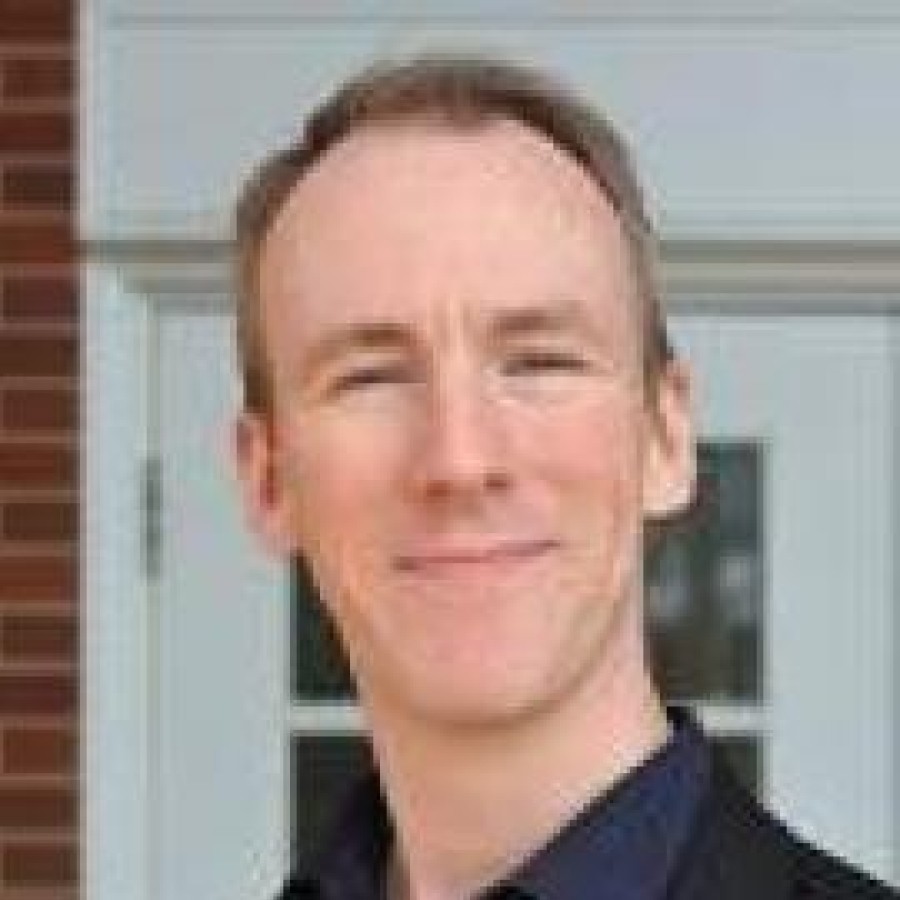 Headshot of Mark Leeming smiling in front of brick wall and door. 