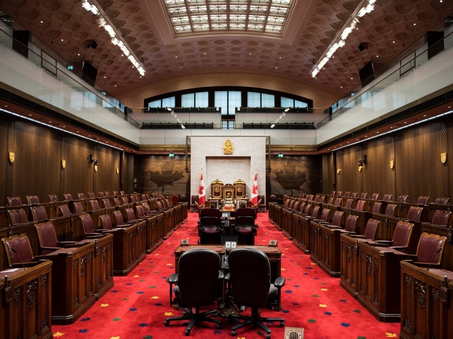 Large room with dark wood paneling and seats along both sides and a central aisle with desks and red carpet.